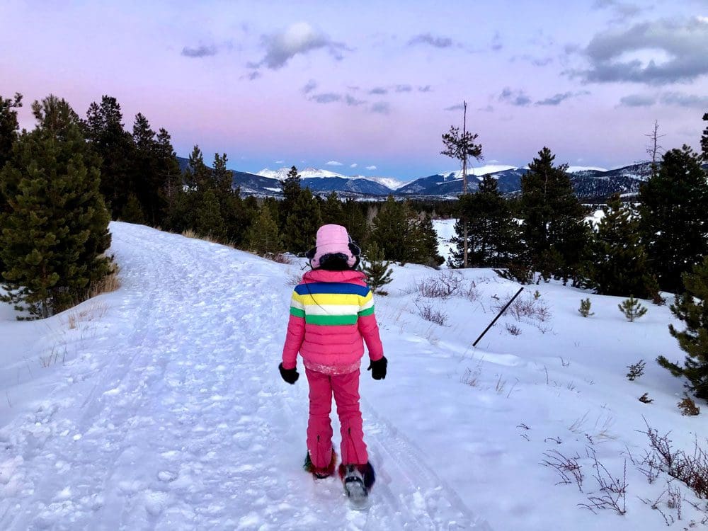 A young girl wearing a pink snowsuit hikes along a wintery trail at sunset while enjoying a family vacation to Breckenridge during the winter with kids.