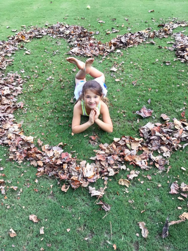 A young girl lays in the middle of a pile of leaves in the shape of a heart.
