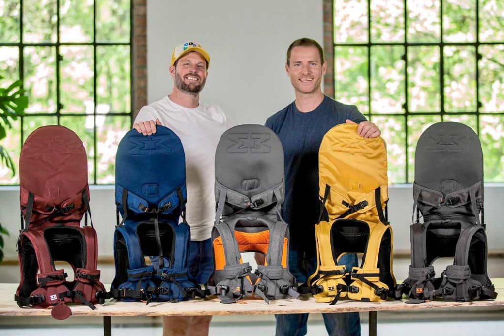 Two men stand behind a table showing five MiniMeis G4 Shoulder Carrier in various colors, including red, yellow, and blue.
