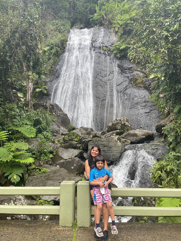 A mom holds her young son, while leaning against a fence, behind them in a watterfall.