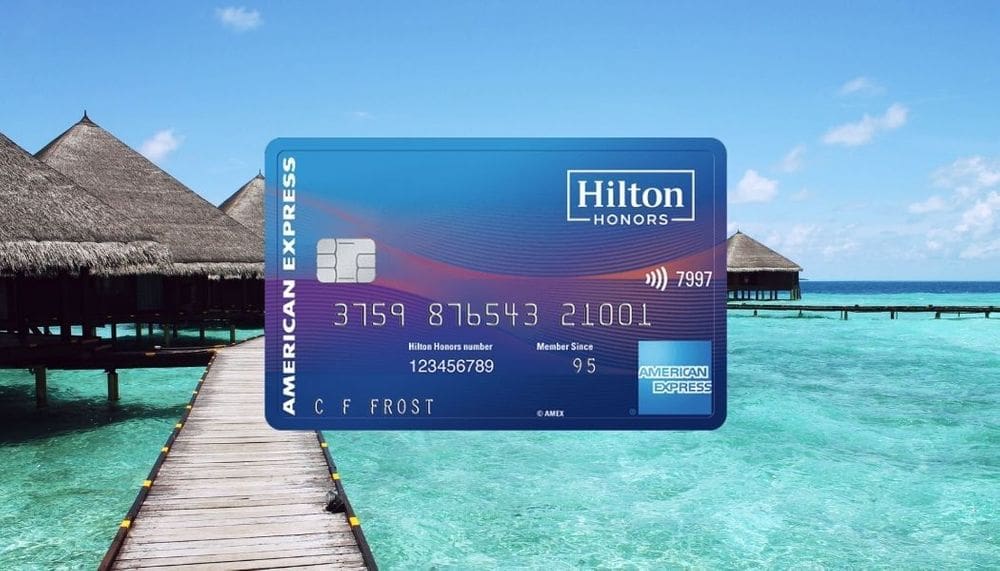 A close up of the American Express Hilton Honors Surpass credit card, nestled over an image of a boardwalk water overwater bungalows and turquoise waters.
