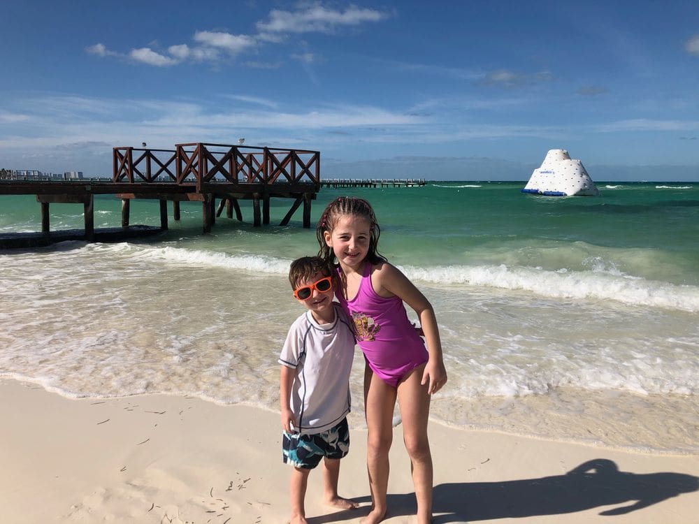 Two kids in bathing suits stand together on the beach at Hyatt Ziva in Cancun, with an elevated dock behind them.