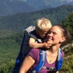 A mom carries her baby on her back using a backpack-style carrier, the baby reaches up toward the mother to give her a snuggle, while hiking in the mountains.