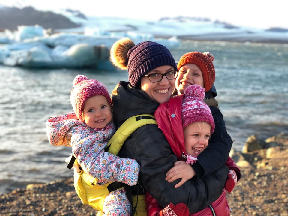 A mom holds her baby on her back with a carrier, while hugging her other two children two her chest while exploring Iceland together.