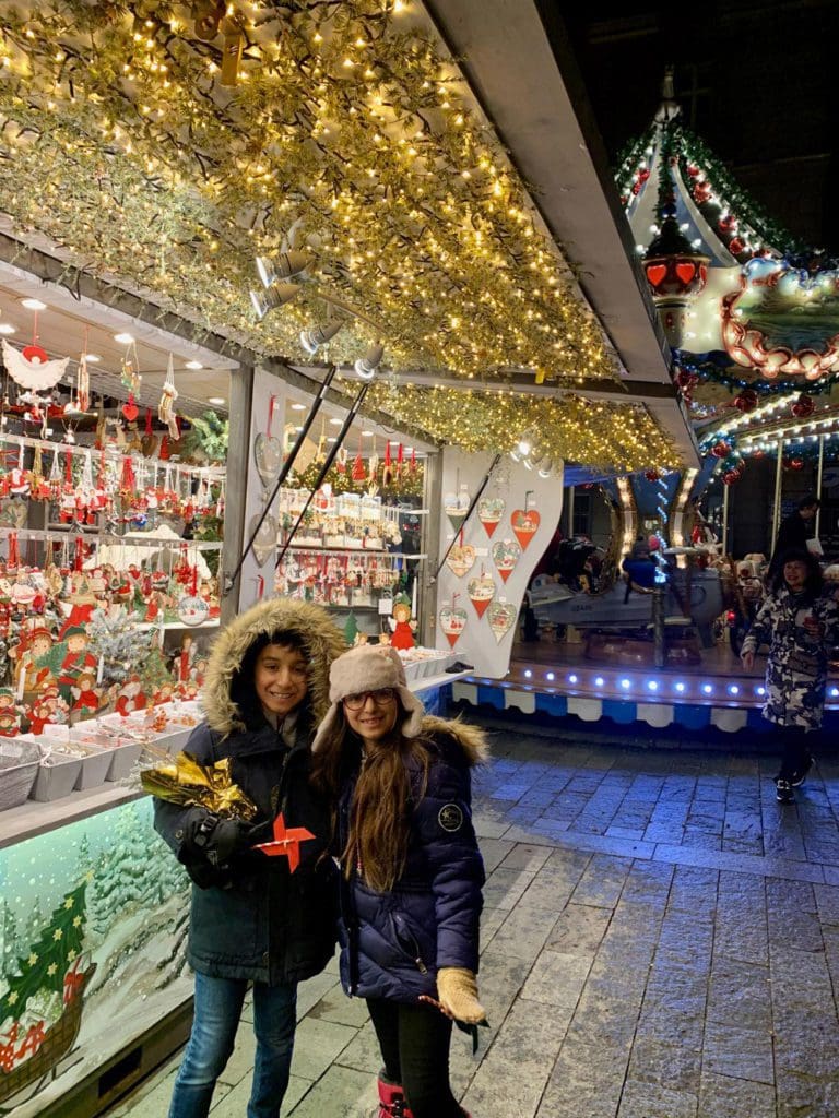 Two kids stand together in winter gear at a European Christmas market.