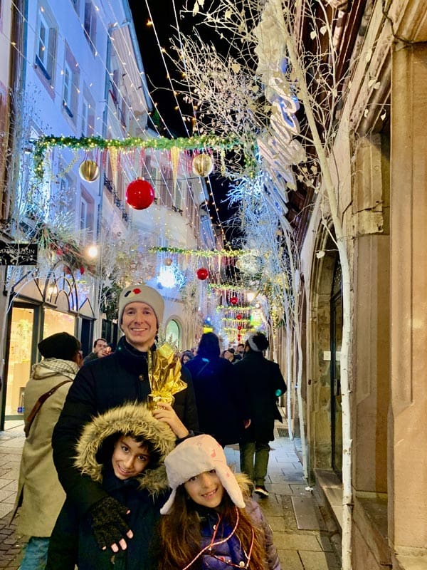 Two kids stand with their dad while wandering a Chrismtas-lined street at the Strasbourg Christmas Market.