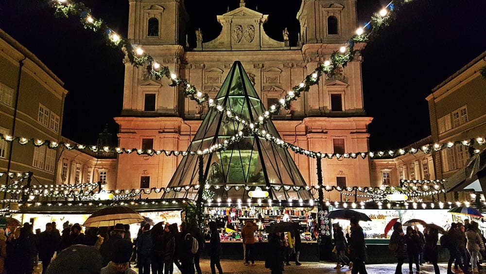 People wander around the Salzburg Christmas Market, the center of which holds a tall glass Christmas tree and twinkle lights.