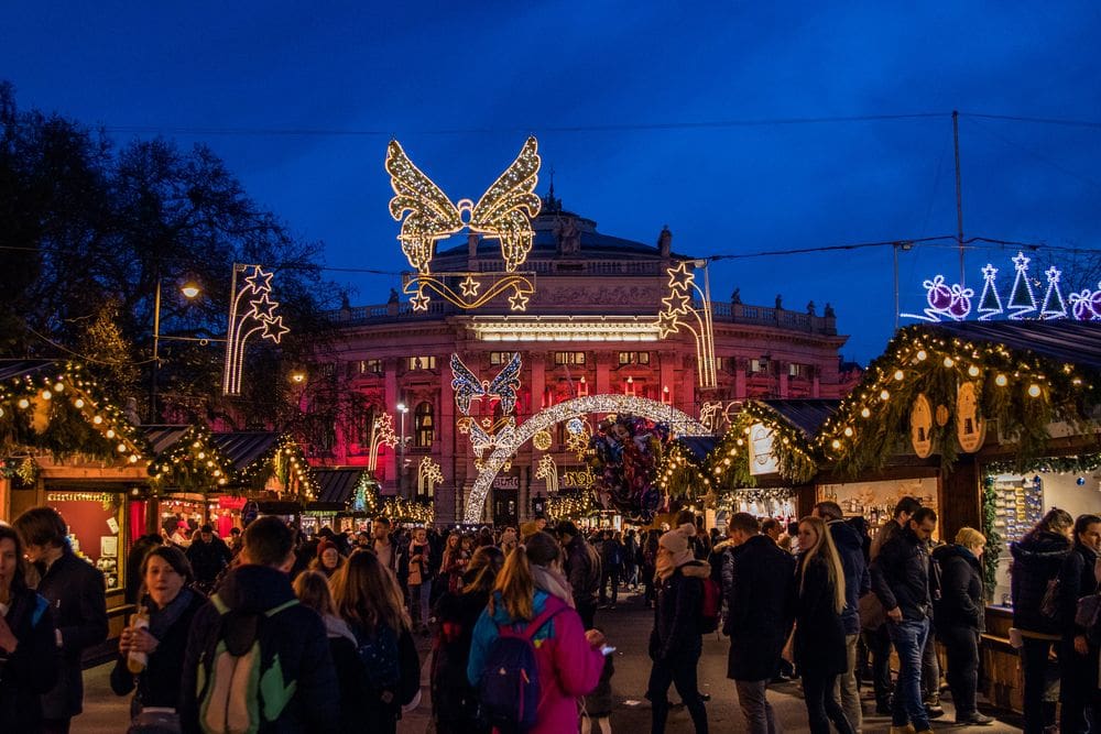 A crowd of people explores the Christmas market in Vienna, well-lit with Christmas lights at night.