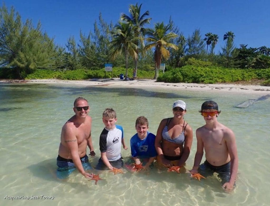 A family of five stands in the water, each family member is holding a starfish, while visiting the Cayman Islands.