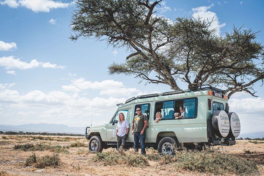 Parents stand near a safari jeep, while their kids sit inside, during a safari in Tanzania.