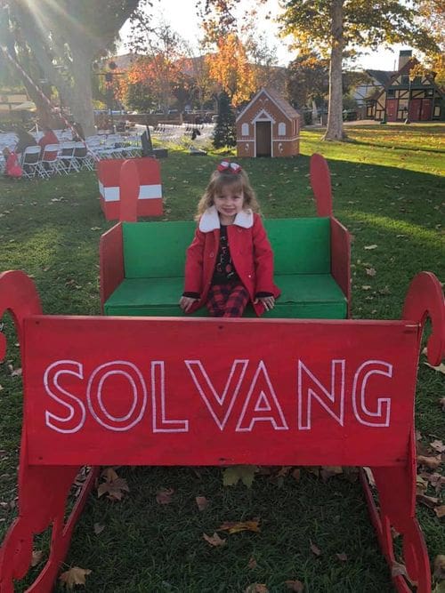 A young girl, dressed in red, sits in a red sleigh reading "SOLVANG" while celebrating the holidays with her family.