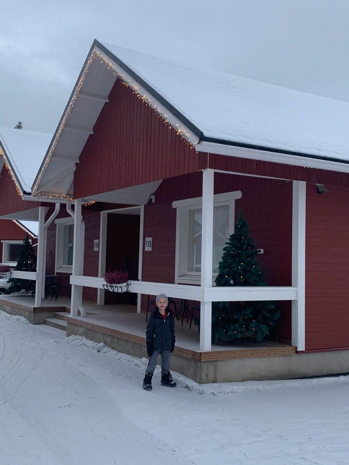 A young boy stands in front of a red cabin at Santa's Village in Lapland, a great accommodation option on our Finland winter itinerary for families.
