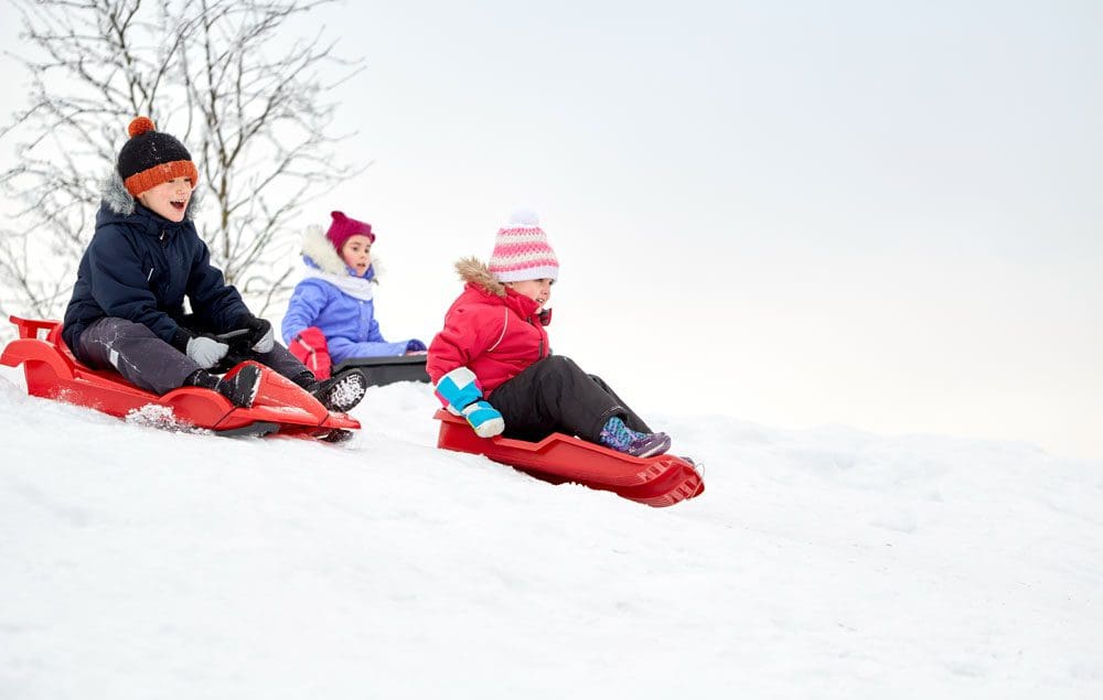 Three kids sled down a snowy hill, all wearing winter gear, including colorful hats.