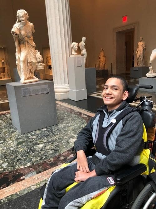 A teen boy, who uses a wheel chair, is sitting next to a sculpture at the Met in NYC, one of the best vacation spots in the US to impress teens and tweens.