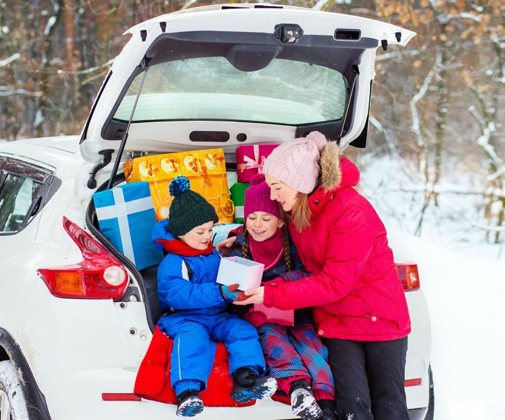 A mom hands her two children a gift in the back of a trunk laden with holiday gifts on a winter's day.