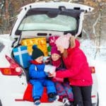 A mom hands her two children a gift in the back of a trunk laden with holiday gifts on a winter's day.