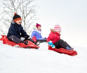 Three kids sled down a snowy hill, all wearing winter gear, including colorful hats.