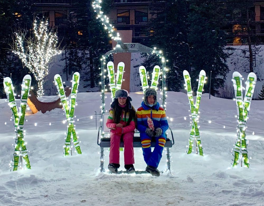Two kids sit near holiday lights that look like skis in Vail, one of the best places for a White Christmas in the United States for families.