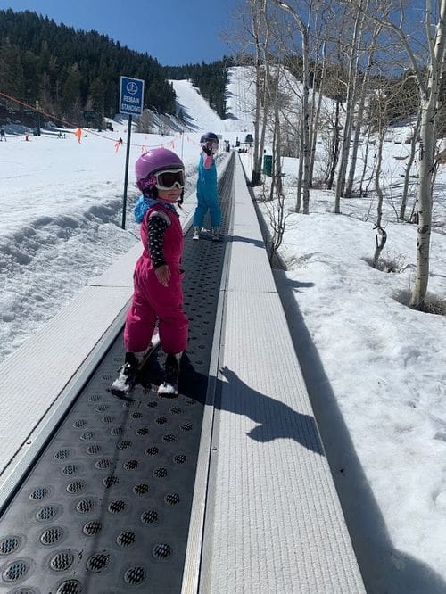 Two young kids in full snow gear ride the magic carpet up a s