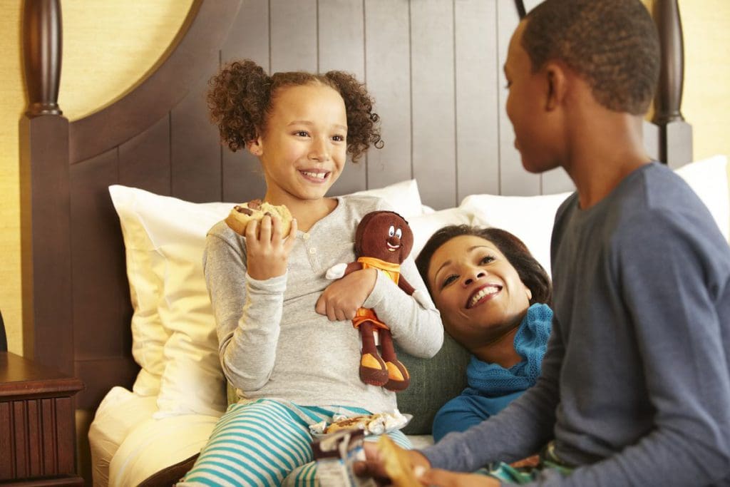 A mom of color and her two kids sit on a bed, enjoying their stay at the Hershey Lodge.