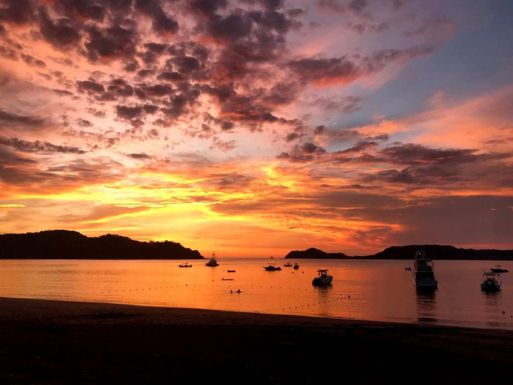 A brilliant sunset in Costa Rica, featuring hues of yellow, pink, and orange, with several boat on the water at dusk.