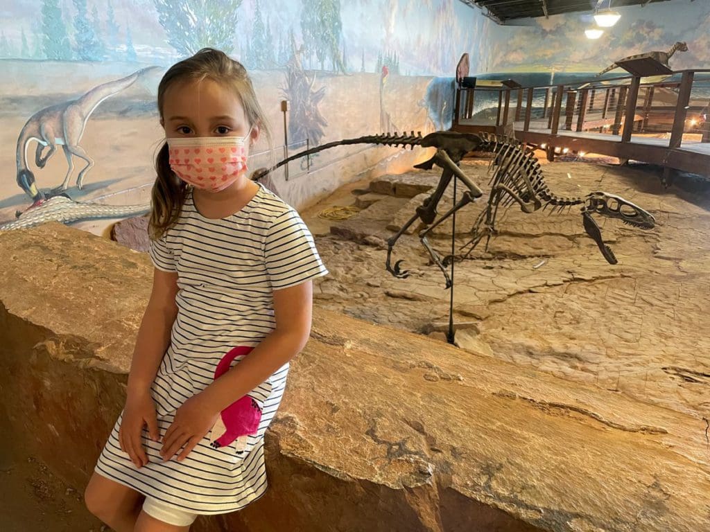 A young girl sits near an exhibit at the Dinosaur National Monument in Utah, with a small dinosaur skeleton behind her.