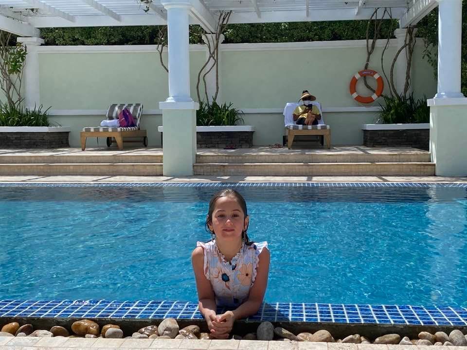 A young girl in a pool leans over the ledge of the pool and smiles.