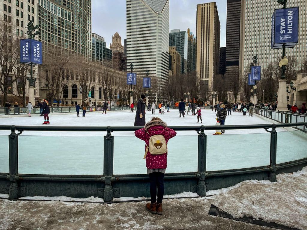 A young girl looks over a skating rink barrier at several skaters enjoying a winter day at Millennium Park in Chicago, one of the best places to visit with your young daughter in America.
