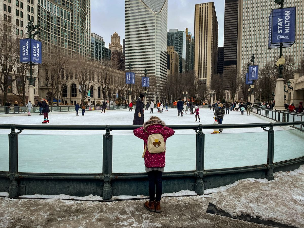 A young girl looks over a skating rink barrier at several skaters enjoying a winter day at Millennium Park, one of the best kids activities in Chicago.