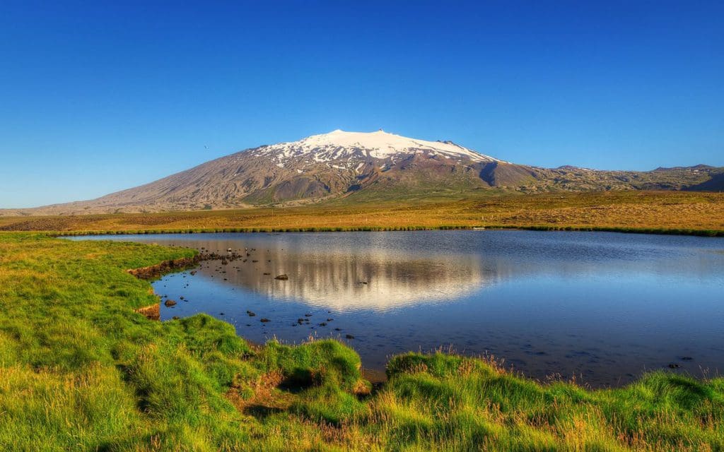 A serene view of Snaefellsjokull National Park, featuring a snow-topped mountain, glacial lake, and greenery.