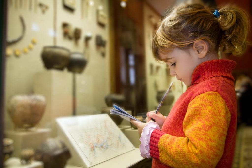 A young girl looks down at a museum exhibit, writing notes.