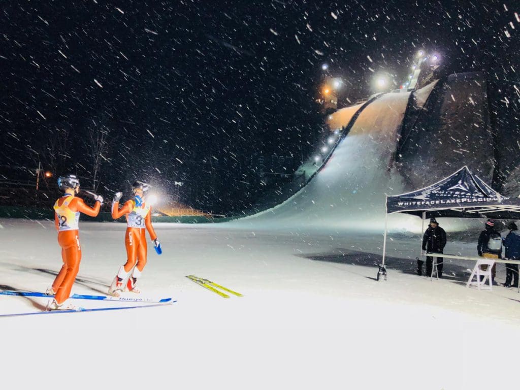 Two skiers stand at the base of the large ski jump near Lake Placid.