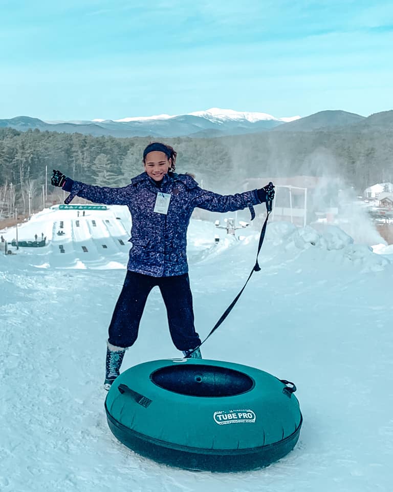 A teenage girl stands with her hands high in the air next to her snow tube, while enjoying a winter day.