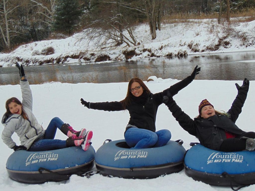 Three women, each sitting on their own snow tube, throw their hands in the air as they enjoy a winter day at Holiday Mountain Ski & Fun Park.