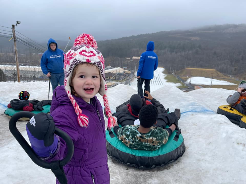 A young girl looks at the camera, as other prepare to snow tube down a hill behind her.