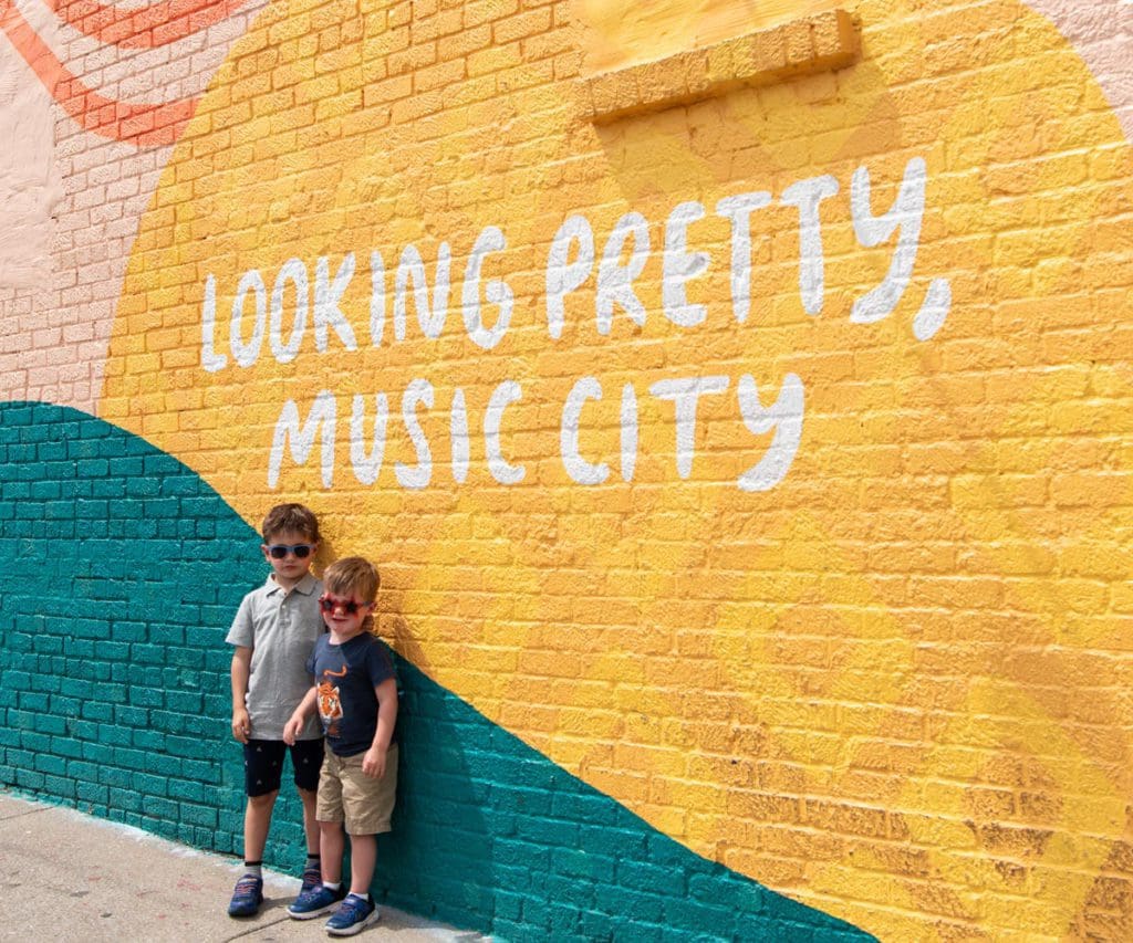 Two boys wearing sunglasses stand together in front of brightly colored street art in Nashville, reading "Looking Pretty, Music City".