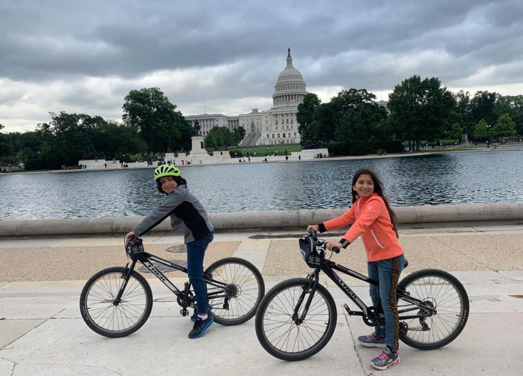 Two kids on bikes rest along the National Mall with monuments in the distance.