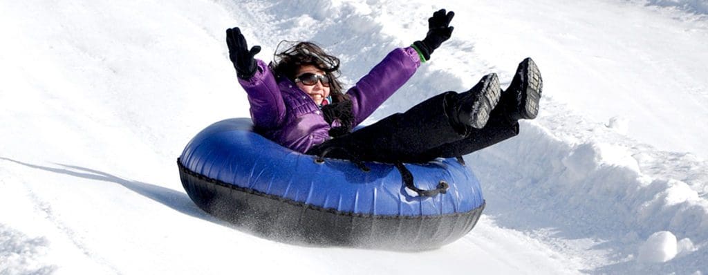 A young girl wearing a purple coat flies down the snow tubing hill at Rocking Horse Ranch, one of the best hotels with snow tubing near NYC for families.