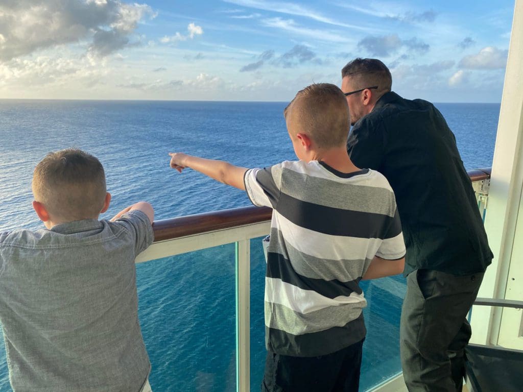 A dad and his two kids enjoy an ocean view from a balcony on a Royal Caribbean Cruise.