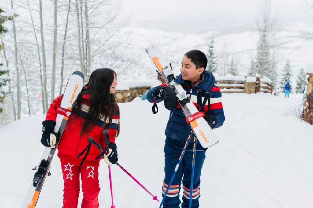 Two kids holding their own skis look at each other and laugh, as they get ready to ski on a beautiful day.