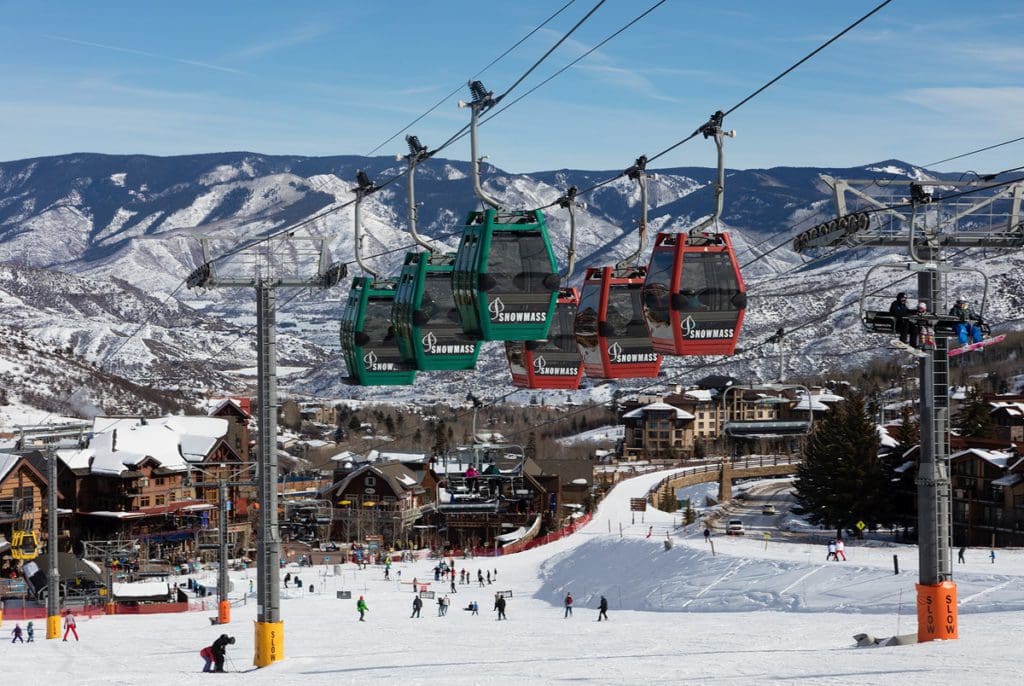 A view of Snowmass Mountain, including several gondolas overhead, skiers on the slopes, and snowy mountains in the distance.