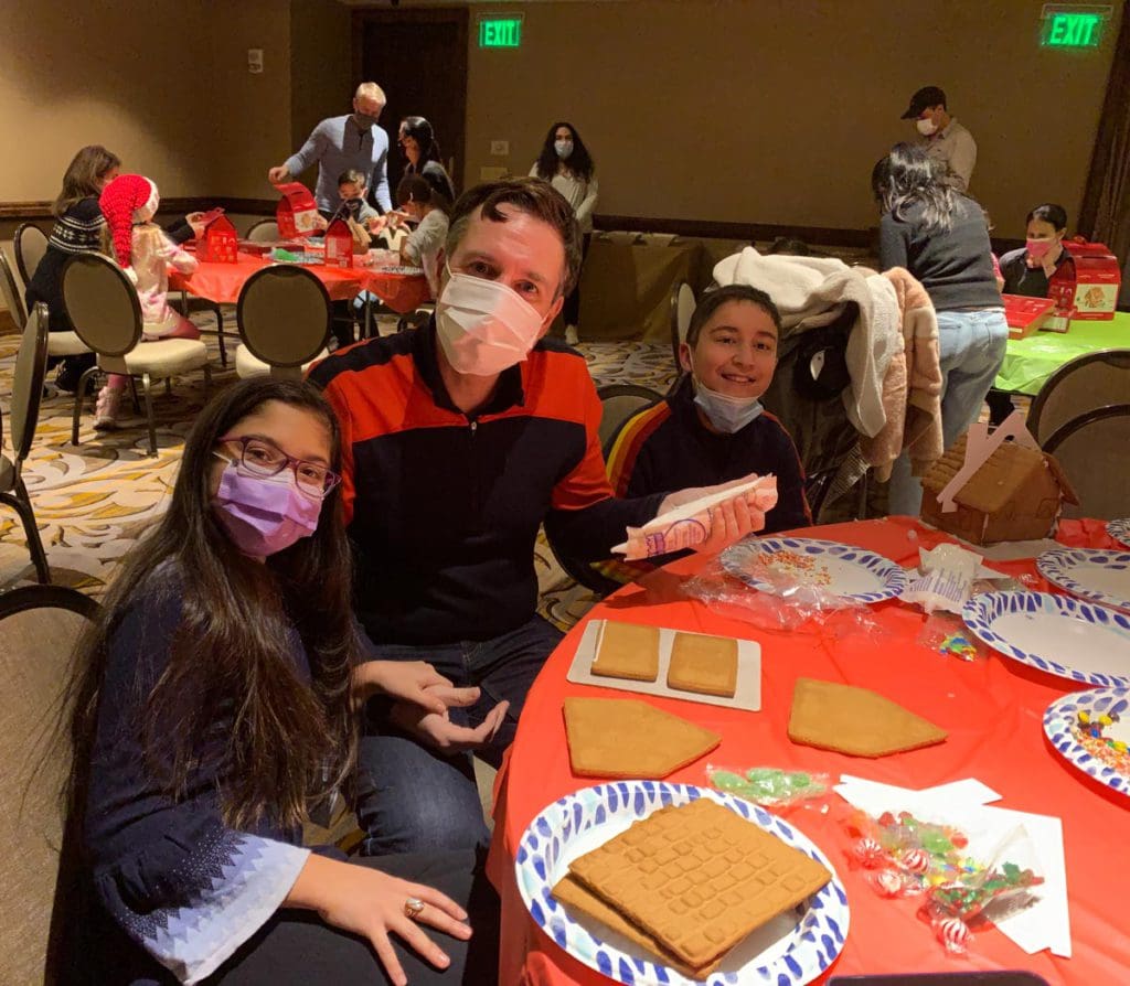 A dad and his two kids sit around a table making a gingerbread house.