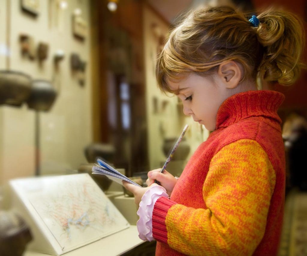 A young girl looks down at a museum exhibit, writing notes in NYC, one of the best places to visit with your young daughter in America.