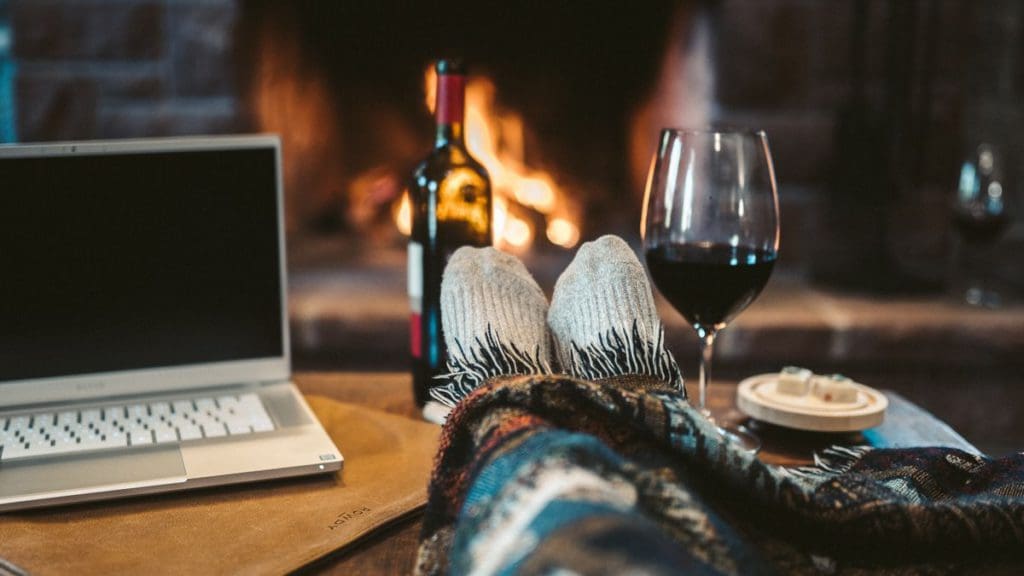 Feet stick out from a blanket with a view of a fireplace, wine, and computer during a family weekend away, one of the best travel gifts for Mothers Day.