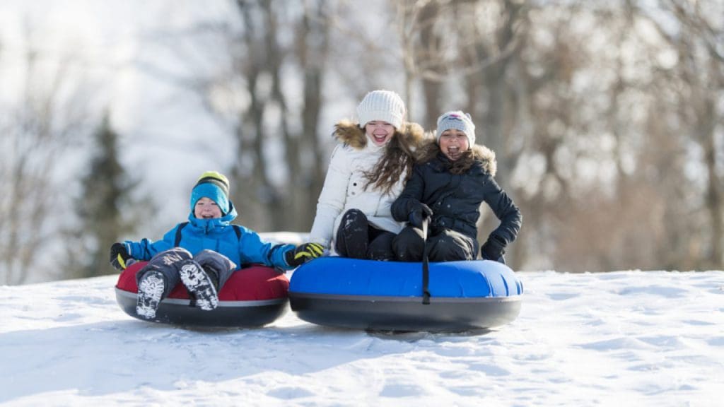A mom stands behind two kids as they snow tube down a winter hill at the Skytop Lodge, one of the best hotels with snow tubing near NYC for families.