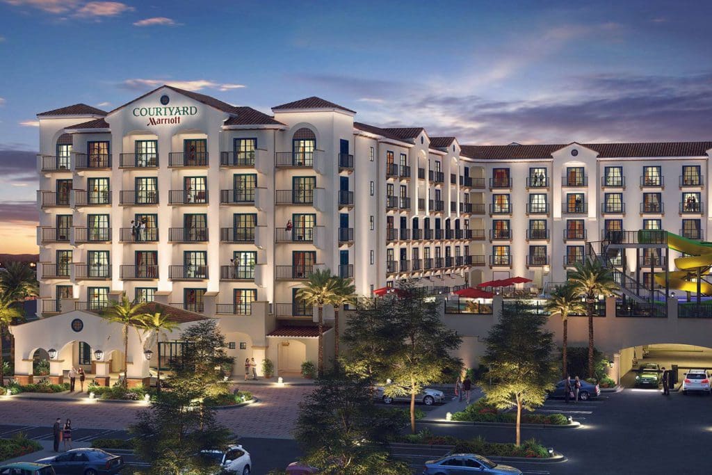 A view of the exterior of the Courtyard by Marriott Anaheim Theme Park Entrance at dusk, featuring talk hotel buildings and palm trees.