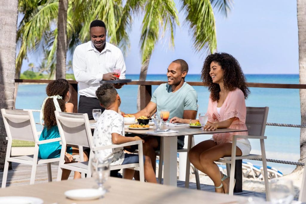 A family of four sits at a dinner table at Hilton Rose Hall Resort & Spa, while a staff member serves them.