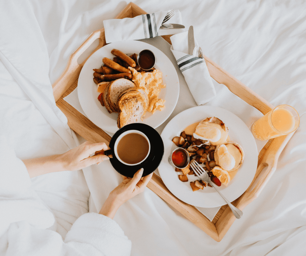 Looking down on outstretched hands holding a coffee above a breakfast plate on a hotel bed, featuring sausage, toast, and eggs. Breakfast is just one of the perks that can come with using a Travel Advisor for a Family Vacation.
