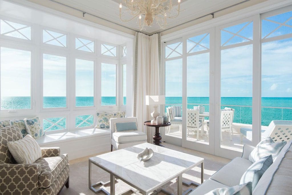 Inside a hotel living room, featuring floor to ceiling windows onto the ocean and white, beach-theme furnishings.