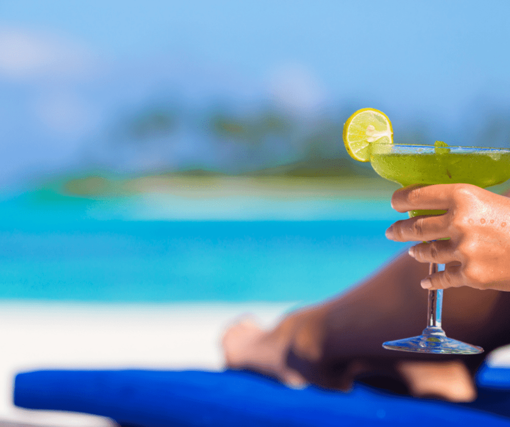 A hand holds a margarita, while the individual sits on a beach.
